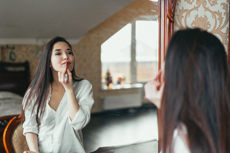 Beautiful woman applying lipstick in front of mirror at home