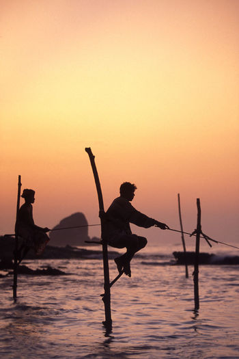 Side view of silhouette man and woman fishing in sea from wooden posts at sunset