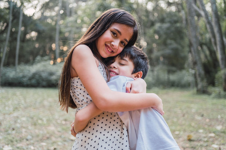 Side view of cheerful girl looking at camera while embracing sibling with closed eyes on lawn