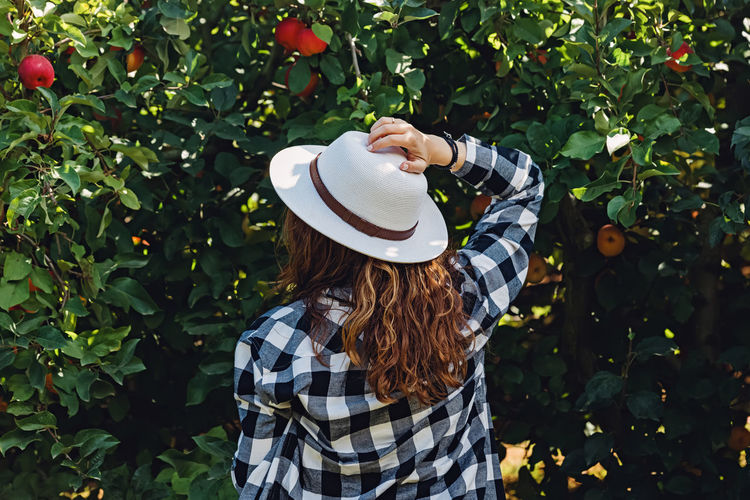 Woman in a hat standing near the tree with red ripe apples, countryside lifestyle person