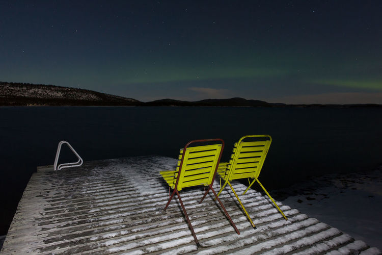Empty chairs on pier by lake against sky at night