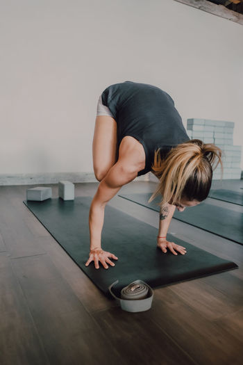 Back view upside down of unrecognizable female athlete in activewear practicing yoga in bakasana position on black sports mat in spacious modern studio