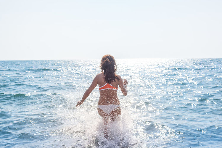 Rear view of woman splashing water in sea against sky during sunny day