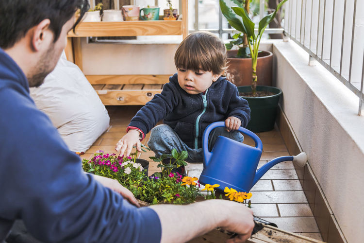 Father planting plant while sitting by boy holding watering can at balcony