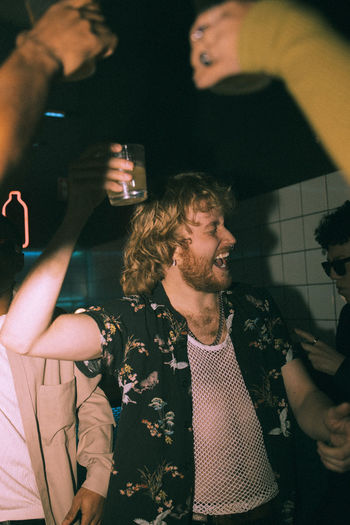 Cheerful young man holding drink dancing with friends at nightclub