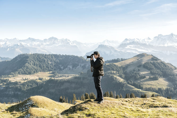 Young man photographing while standing on mountain against sky