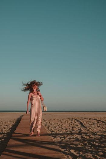 Woman with tousled hair walking on boardwalk at beach against blue sky