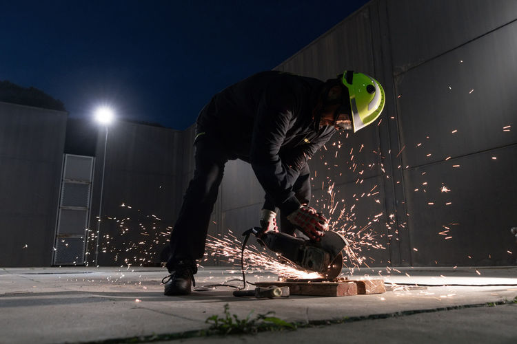 Full body of unrecognizable male welder in protective helmet and goggles cutting iron details with chop saw during work at industrial factory at night