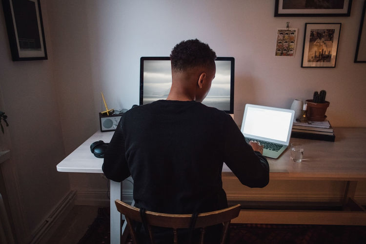 Rear view of young freelance worker using laptop and computer at desk