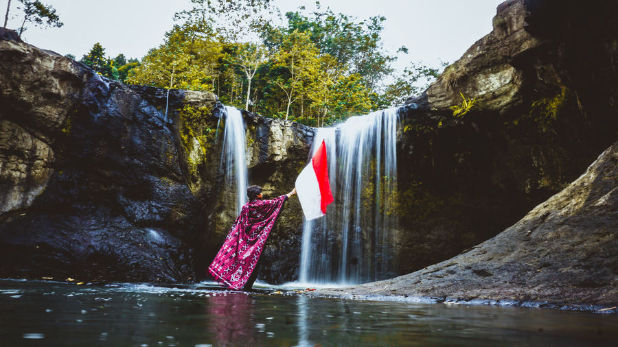 Rear view of woman holding flag while standing by waterfall