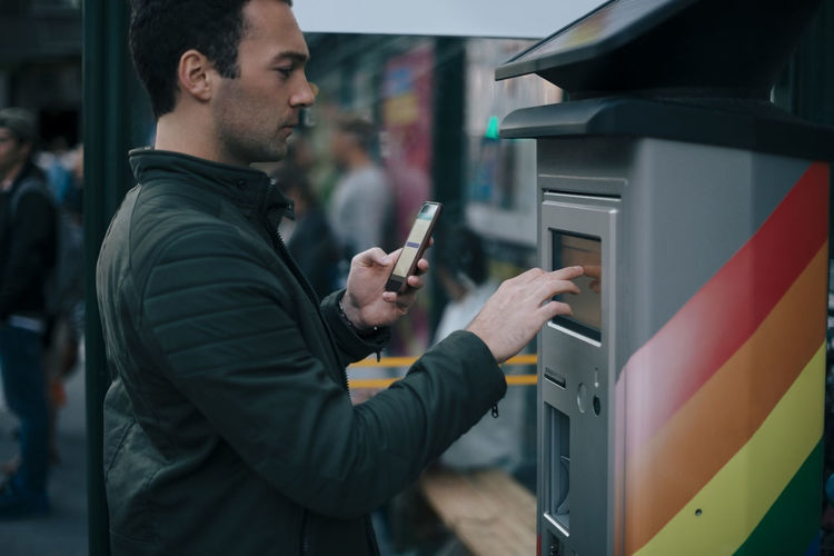 Side view of man touching atm screen while holding smart phone