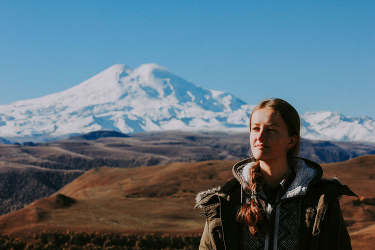 Young woman looking away while standing against snowcapped mountain and sky