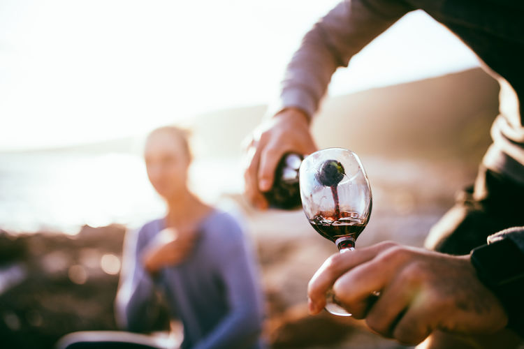 Midsection of man pouring red wine while woman sitting in background during sunset