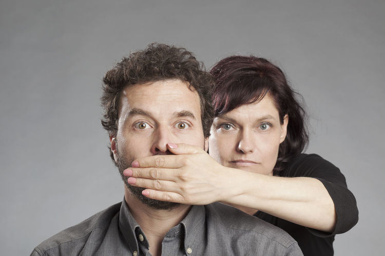 Portrait of young woman covering man mouth against white background