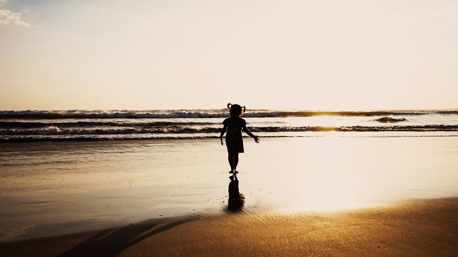 Child standing on beach against sky during sunset