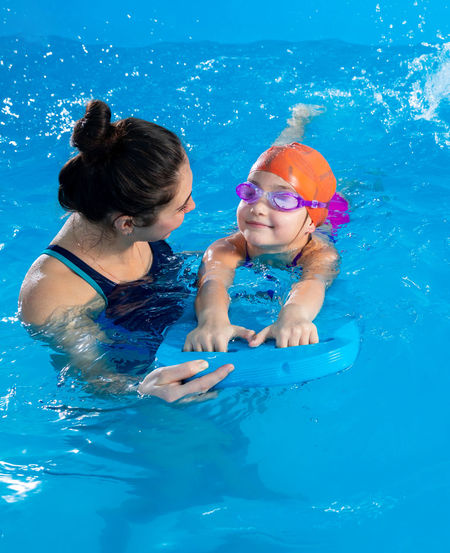 Trainer teaching little girl how to swim in indoor pool with pool floating board