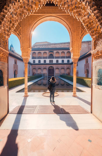 Anonymous woman standing under arch near calm pool and shooting sunlit court of the myrtles of comares palace on weekend day in granada, spain