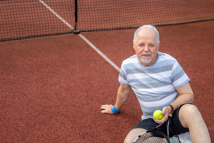 Portrait of smiling man holding tennis ball sitting on court