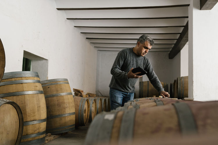 Winemaker holding digital tablet while examining wine barrels at cellar