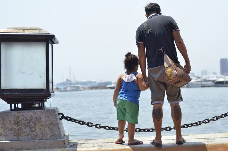 Rear view of father with daughter standing on retaining wall by sea