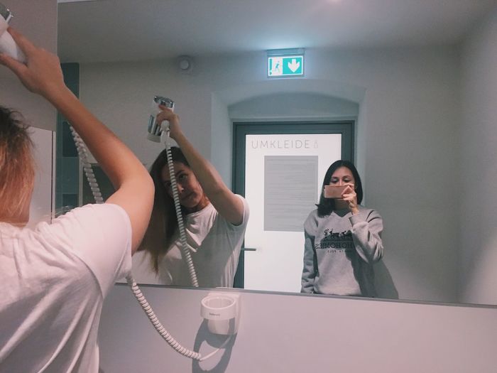 Young woman taking mirror selfie while friend drying hair in restroom