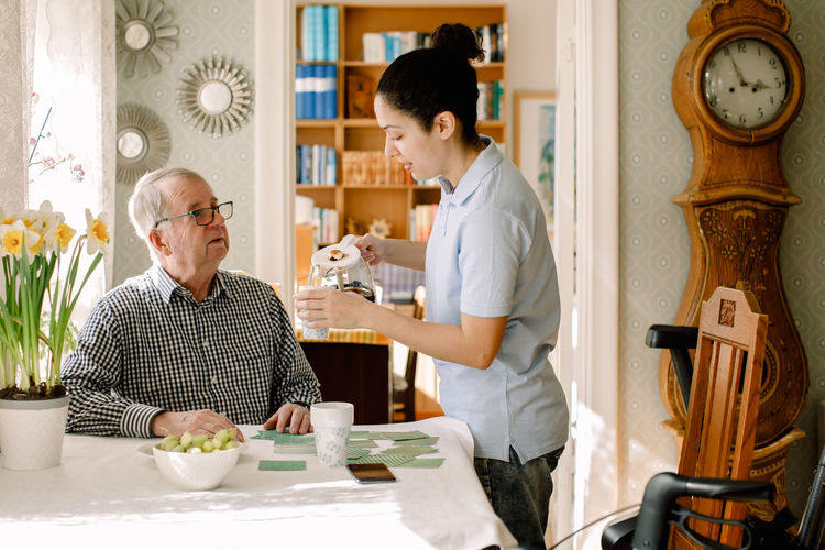 Retired senior man looking at young female caregiver pouring coffee in cup