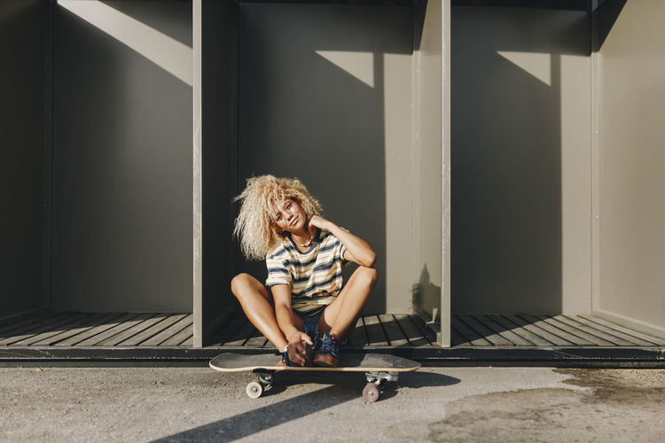 Woman with skateboard sitting against metallic cabins on sunny day