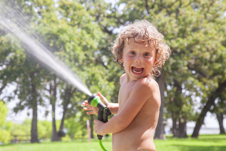 Portrait of shirtless boy holding gardening hose standing against trees