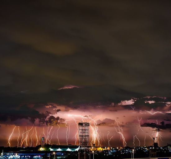 Illuminated buildings in city against sky at night with massive lightning strikes