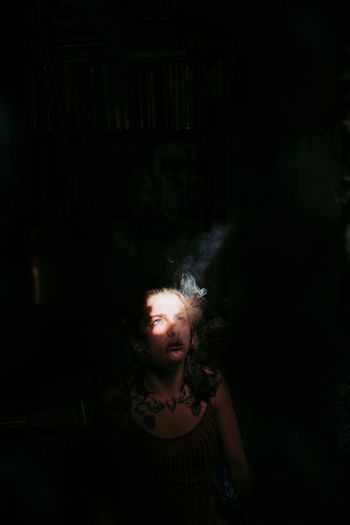 High angle of casual woman standing and looking up while smoking under beam of light in dark room