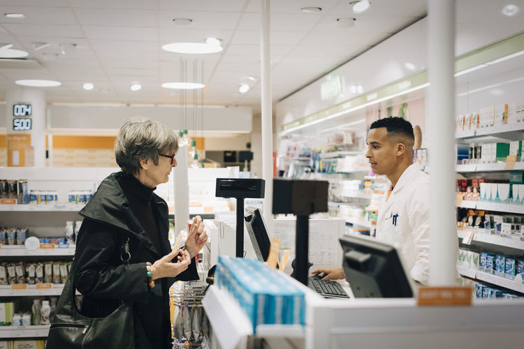 Side view of senior female customer talking with male pharmacist at checkout in store