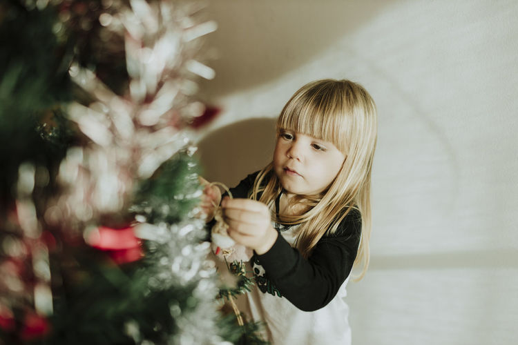 Blond girl decorating christmas tree against wall at home