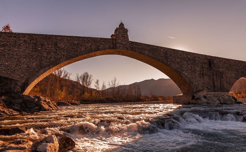 Landscape of a medieval bridge over a turbulent river at sunset in winter
