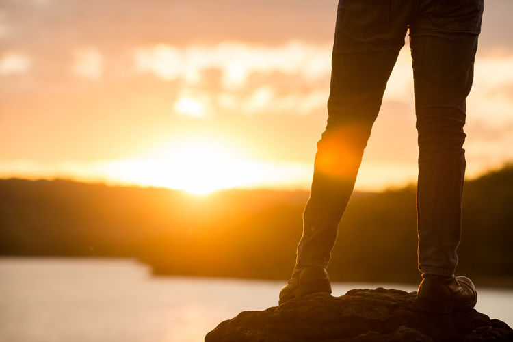 Low section of man standing on rock against sky during sunset