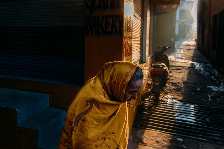 Varanasi, india - february, 2018: side view of old indian female in traditional saffron colored shawl standing next to local bakery on narrow dirty street