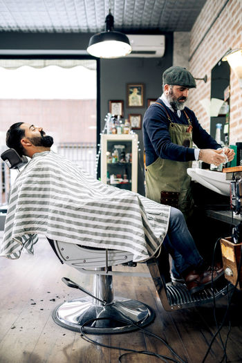Side view of positive young bearded hispanic male client sitting on chair near middle aged barber preparing for haircut in traditional barbershop