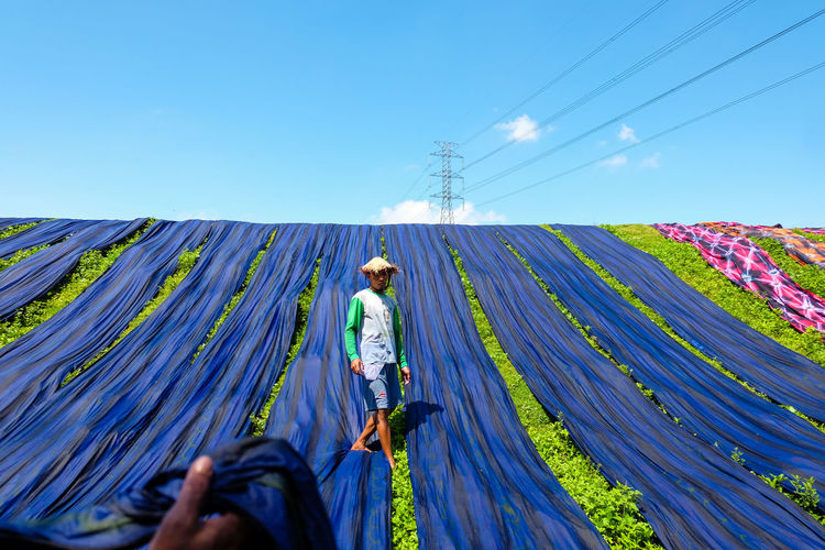 Man drying blue fabrics on grassy field against blue sky during sunny day