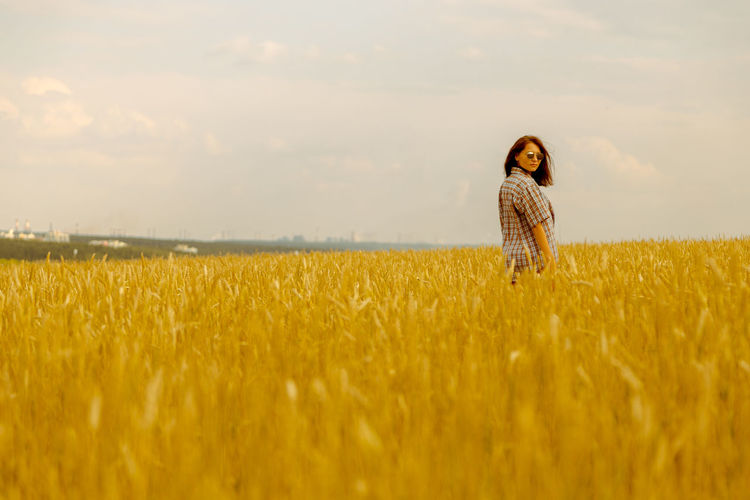 Portrait of young woman standing on wheat field against cloudy sky during sunset