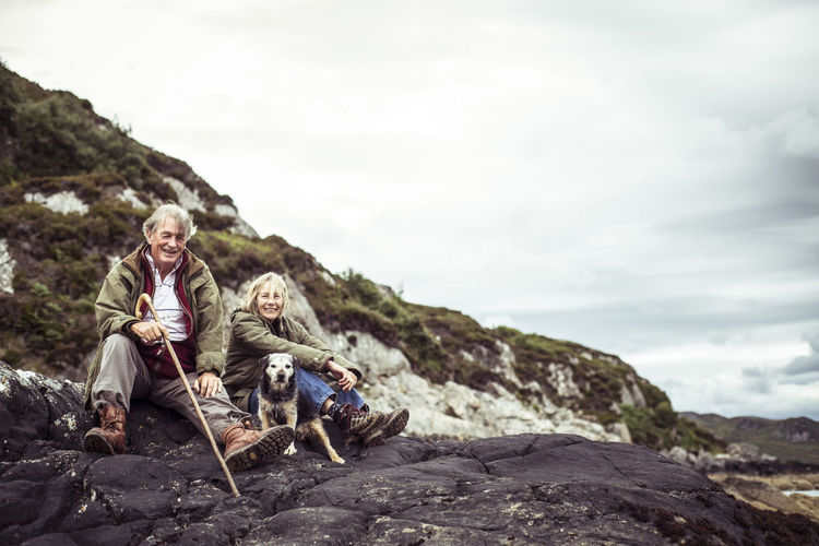 Retired couple sit with dog on rocks by ocean in rugged scotland