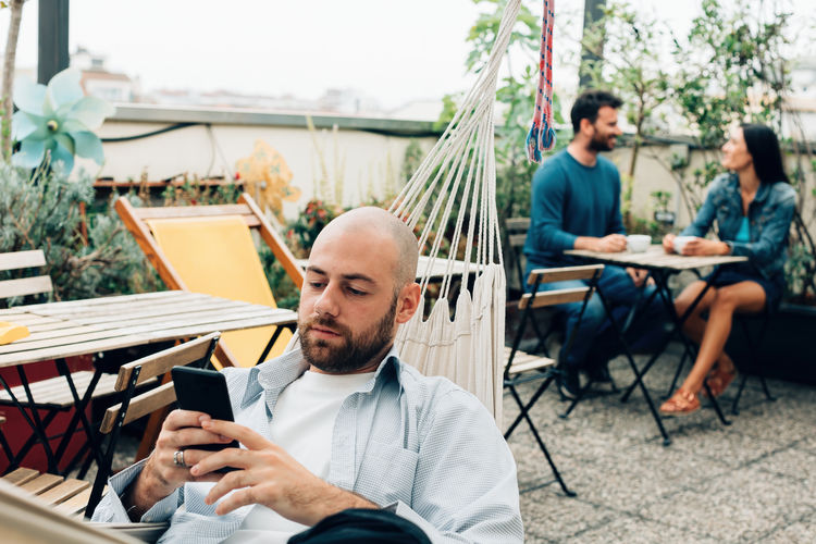 Shaved head boy in the hammock spends time with his mobile phone on an outdoor terrace - hostel bar 