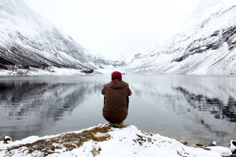 Rear view of man sitting at lakeshore against snow covered mountains