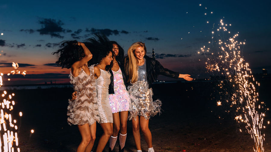 Cheerful young female friends with firework at beach against sky during sunset
