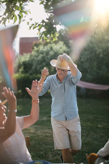 Senior woman clapping while man wearing straw hat and walking in backyard during sunny day