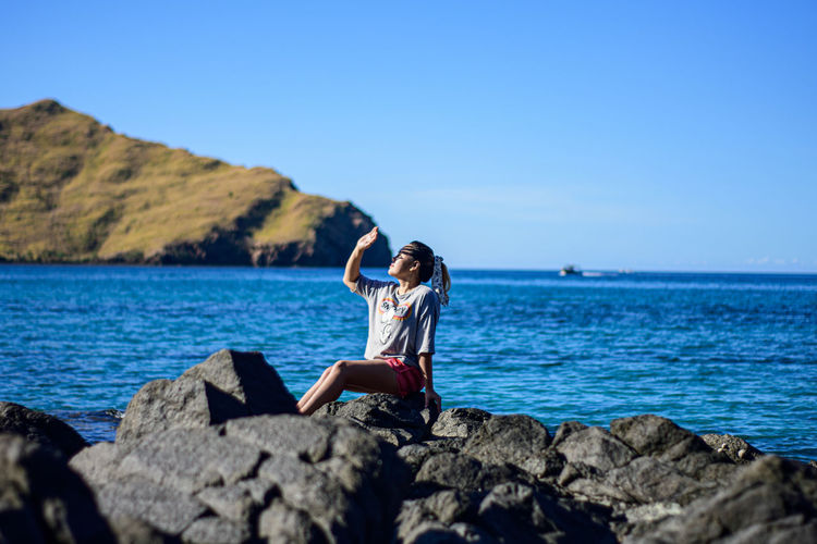 Side view of woman standing on rock by sea against clear blue sky