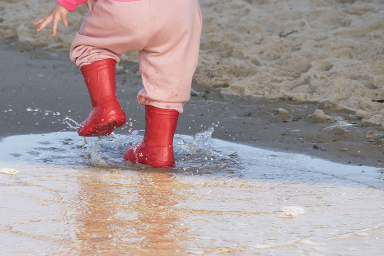 Low section of child wearing rubber boots while stamping feet in water on shore at beach