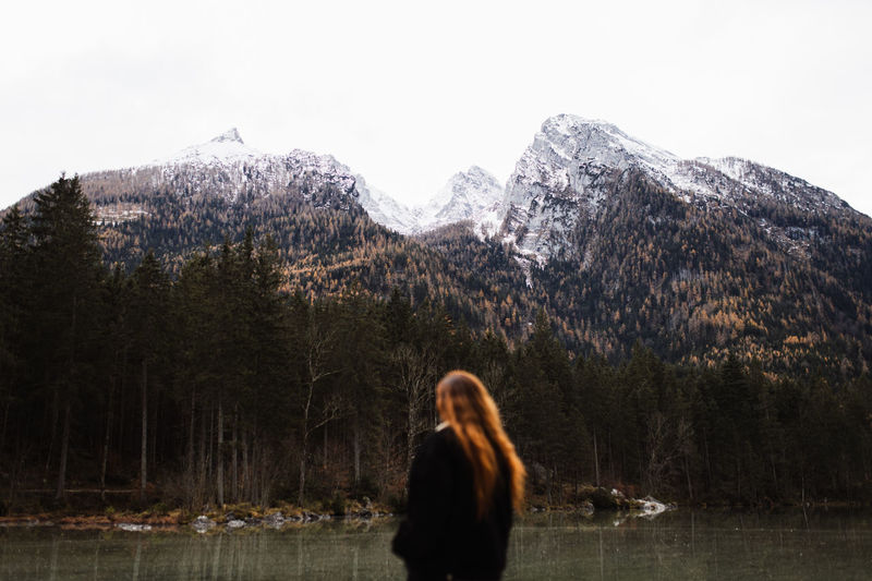 Back view of unrecognizable female tourist enjoying amazing landscape of calm lake and majestic mountains with forested slopes and snowy peaks in cloudy autumn day