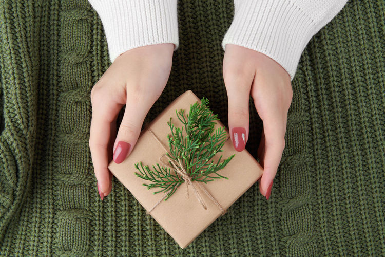Female hands holding a kraft gift with fir branch on knitted green sweater. cozy christmas stilllife