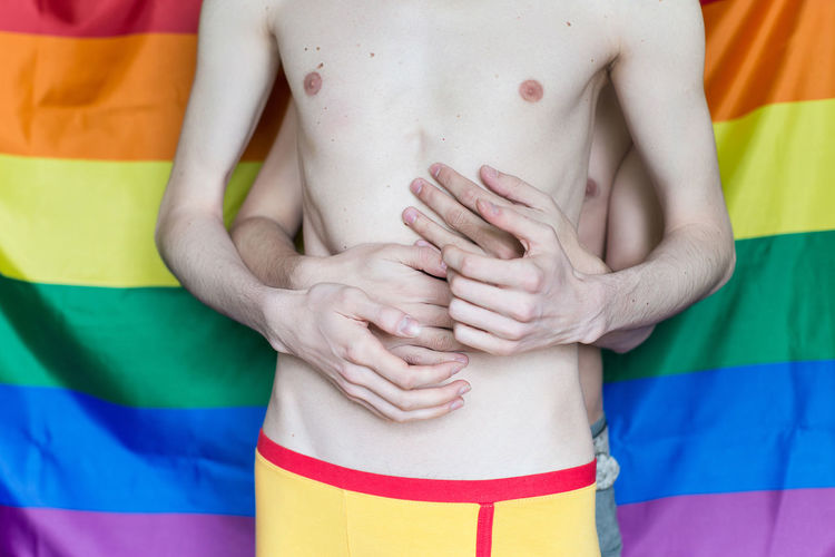 Midsection of shirtless gay couple embracing against rainbow flag