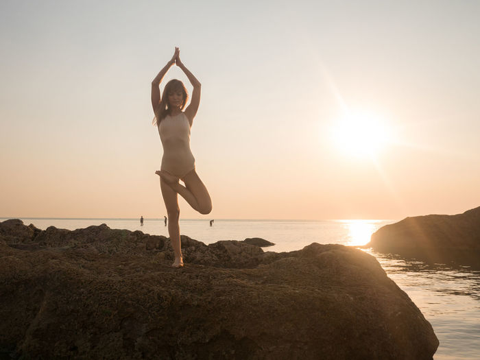 Full length of woman doing tree pose on rock at beach against sky during sunset
