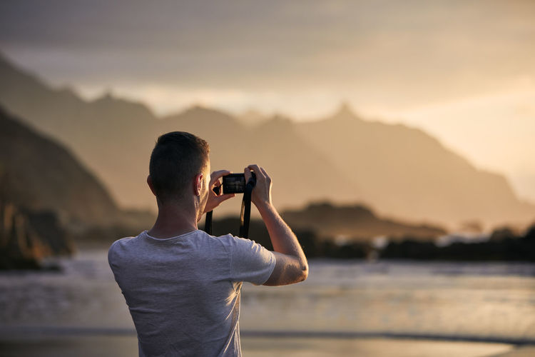 Rear view of man during photographing landscape with cliff. photographer on beach at moody sunset. 